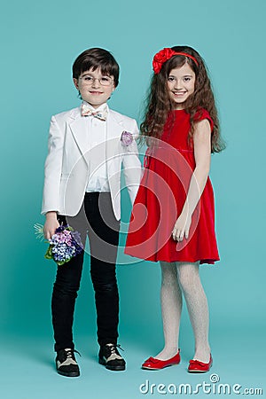 Image of a two kids in elegant clothes, holding a flowers, girl in red dress smiling, isolated on a blue background. Stock Photo