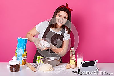 Image of woman making and preparing pastry in bakery kitchen. Adding flour bits. Female has pleasant facial expession, looks Stock Photo