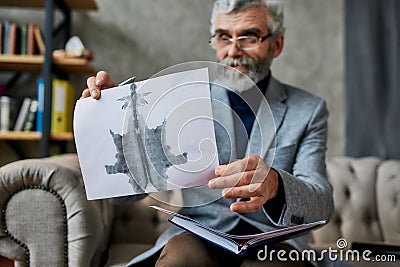 Imagine. Mature psychologist holding picture with ink stain, Rorschach Inkblot during therapy session. Soldier suffering Stock Photo