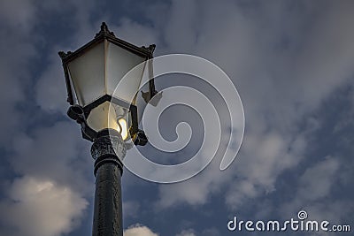 An incandescent bulb with a broken glass glowing in an antique street lamp post.The medieval european lamp post is made of cast st Stock Photo