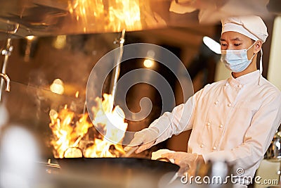 Skillful chef in face mask holding wok in flames Stock Photo