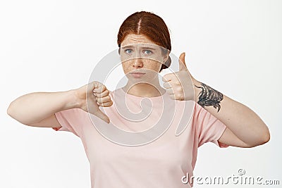 Indecisive redhead girl showing thumbs up, thumb down and look perplexed, judging average not bad result, hesitating to Stock Photo