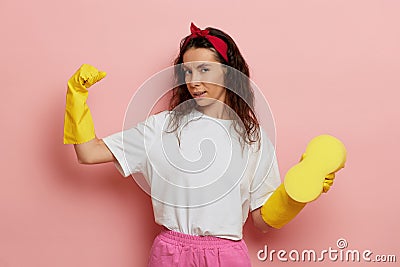 Independent strong housewife shows her muscle, wears yellow gloves and holds a sponge, isolated over pink wall Stock Photo