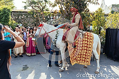 Indian groom ridding white horse with yellow and red pattern fabric, flower necklace and red turban with guests. Editorial Stock Photo