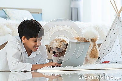 Indian handsome teenage boy lying on floor using laptop computer, relaxing playing with shih tzu dog puppy in bedroom at home. Pet Stock Photo