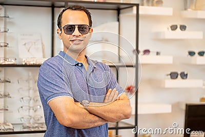 Indian man wear sunglasses with action of arm-crossed of confident in front of shelves with many types of glasses in optical shop Stock Photo