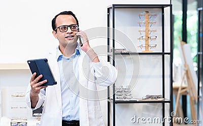 Indian middle aged ophthalmologist or optometrist man work at optometry clinic, using tablet and mobile phone appointing patient Stock Photo