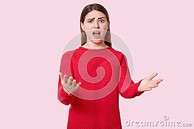 Indignant beautiful young woman gestures with puzzlement, opens mouth widely, raises eyebrows, dressed in red clothes, isolated Stock Photo