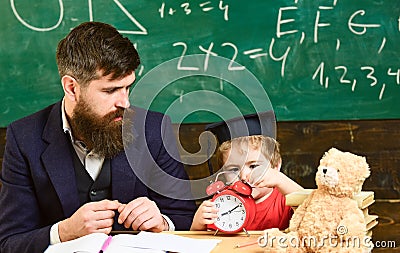 Individual lesson concept. Boy, child on calm face holds alarm clock while teacher talk to kid. Teacher with beard Stock Photo