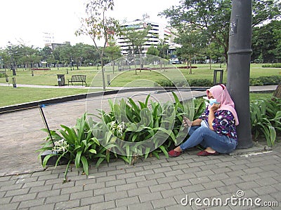 Indonesian woman playing cellphone and sitting on the floor alone in Bandung city park Stock Photo