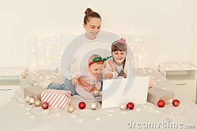 Indoor csot of Caucasian mother with her little daughters, sitting on bed in front of portable computer screen, posing on bed with Stock Photo