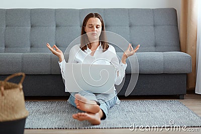 Indoor shot of attractive woman wearing white shirt and jeans sitting on floor and working on laptop, being tired, trying to relax Stock Photo