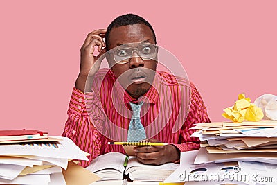 Indoor shot of black employee gazes with stupefaction, has widely opened eyes, wears spectacles, formal pink shirt Stock Photo