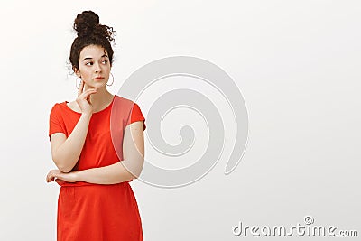 Indoor shot of curious serious female with curly hair combed in bun, wearing red dress, touching face and gazing right Stock Photo