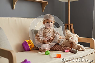 Indoor shot of cute baby girl playing with plush toys and plastic sorter, sitting on sofa alone, little kid with educational toy. Stock Photo