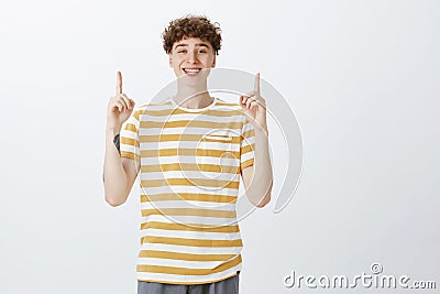 Indoor shot of delighted bright and happy pale guy with curly hairstyle and tattoos in striped yellow t-shirt raising Stock Photo