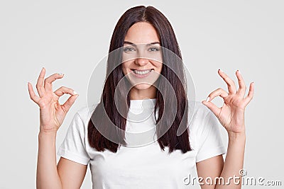 Indoor shot of happy attractive woman with long straight dark hair, makes okay sign with both hands, shows approval, dressed in ca Stock Photo