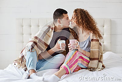 Indoor shot of happy female and male have romantic relationships, drink coffee or tea, covered with plaid in bedroom. Romantic Stock Photo