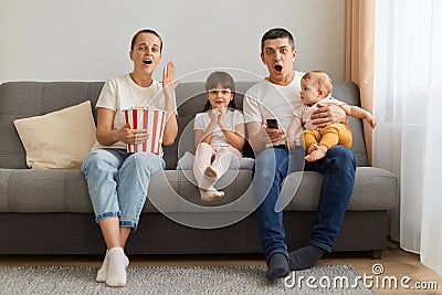 Indoor shot of impressed couple with children sitting on sofa at home in living room and watching shoking tv show or scary movie, Stock Photo
