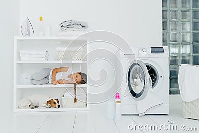 Indoor shot of little girl has sleep on console with favourite dog, has rest in laundry room with washing machine filled of linen Stock Photo