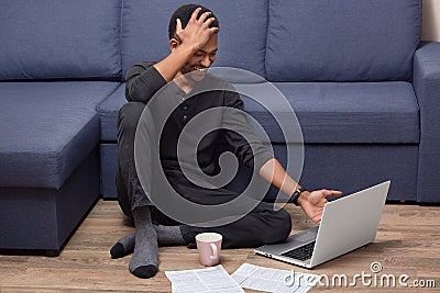 Indoor shot of overjoyed dark skinned man giggles as watches funny video on portable laptop computer, drinks coffee, surrounded Stock Photo