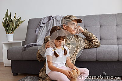 Indoor shot of pensive Caucasian soldier man wearing camouflage uniform returning home after army, spending time with his little Stock Photo