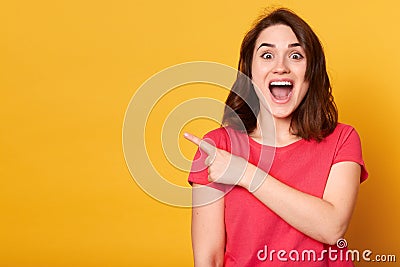 Indoor shot of positive attractive female with dark hair, dressed casually, points aside with index finger, has cheerful Stock Photo