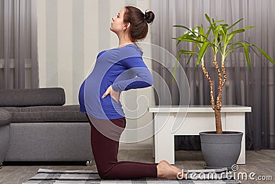 Indoor shot of pregnant lady with hair bun, has big belly, keeps hands on back, dressed in casual clothing, stands on knees, Stock Photo