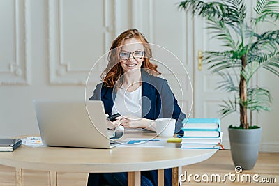 Indoor shot of red haired female economist sends text message to colleague, holds contemporary smart phone, poses at workplace, Stock Photo