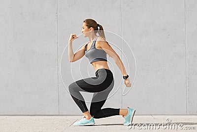 Indoor shot of self determined woman with pony tail, stands on one knee, warms up before cardio training, wears tanktop, leggings, Stock Photo