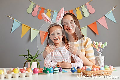 Indoor shot of smiling happy mother and her charming dark haired daughter posing among painted Easter eggs, child wearing bunny Stock Photo