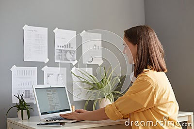 Indoor shot of smiling happy woman with brown hair wearing yellow shirt posing in office, working on portable computer, typing on Stock Photo