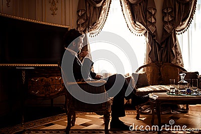 Indoor shot of thoughtful gorgeous businessman or entrepreneur wears black suit, sits in cozy room with luxurious Stock Photo