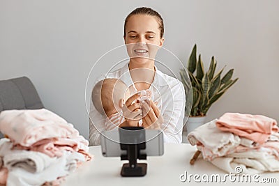 Indoor shot of young adult woman posing with infant daughter in front of tripod with smartphone, filming video for her vlog, Stock Photo