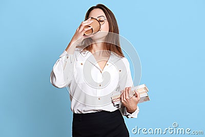 Indoor studio shot of slender hard working office worker having coffee break, drinking hot drink from papercup, holding bunch of Stock Photo