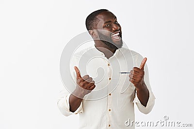 Indor shot of funny and emotive joyful dark-skinned guy with beard in trendy outfit, showing thumbs up and turning face Stock Photo