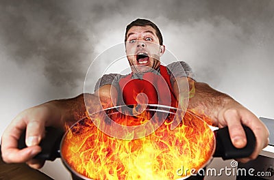 Inexperienced home cook with apron holding pot burning in flames with stress panic face expression Stock Photo