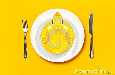 Inflatable mini yellow chicken or duckling on white plate, fork knife on yellow background. Creative food concept, tobacco chicken Stock Photo