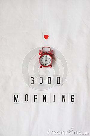 Inscription Good morning, red analog clock, small heart on white rumpled sheets. Top view, flat lay. Vertical. Concept of rest, Stock Photo