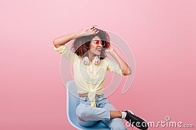 Inspired slim girl in vintage yellow shirt stretching on chair. Indoor portrait of lovely curly african lady in black Stock Photo