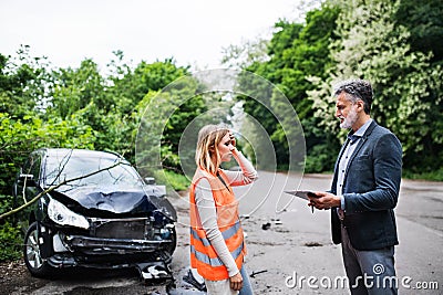 An insurance agent talking to a woman driver by the car on the road after an accident. Stock Photo