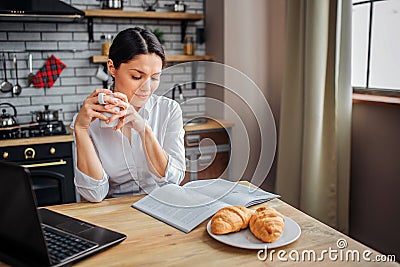 Intelligent adult woman sit at table in kitchen. She read journal and hold white cup with hot drink. Woman look down Stock Photo