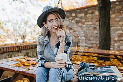 Interested young lady with cup of coffee posing in park in october day. Adorable woman in hat with black manicure Stock Photo