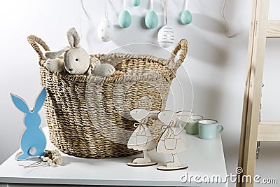 Interior decoration for Easter. rag rabbit peeks out of a wicker willow basket on the table. A garland of wooden painted eggs Stock Photo