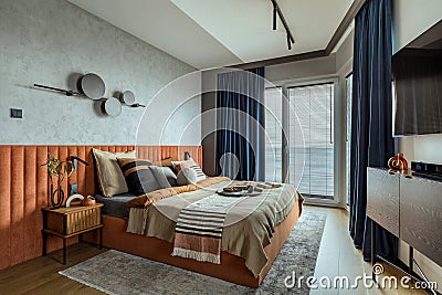 Interior design of elegant bedroom with big orange bed, beige and grey bedclothes, blue curtain, rug, modern lamp, night stand, Stock Photo