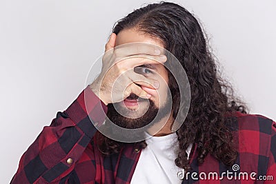 Intrigued man peeks through fingers, covers face with palms, hides himself, afraids of something. Stock Photo