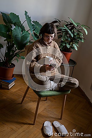 Teen girl sitting on chair with cup of tea, reading news, scrolling photos, messaging in smartphone Stock Photo
