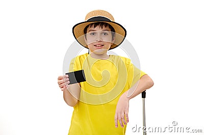 Happy teenage traveler boy in yellow t-shirt and straw hat, with suitcase, showing a blank black plastic card on camera Stock Photo