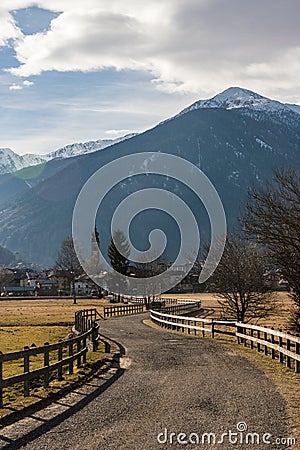 Italian Alps, asphalt road fenced with a wooden fence leading to alpine village Stock Photo