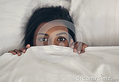 Its too cold to leave bed today. Portrait of a playful young woman hiding under the covers in a bed. Stock Photo